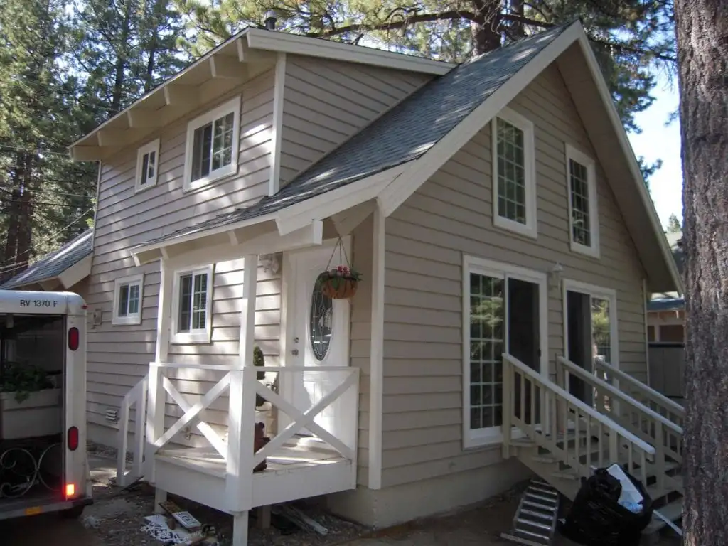 A two-story beige house with fiber cement siding, white trim, and a small porch, surrounded by tall pine trees. The home features large windows and a sliding glass door, with a set of wooden stairs leading to the entrance. Renovation work by 3 Generations Improvements, Folsom.