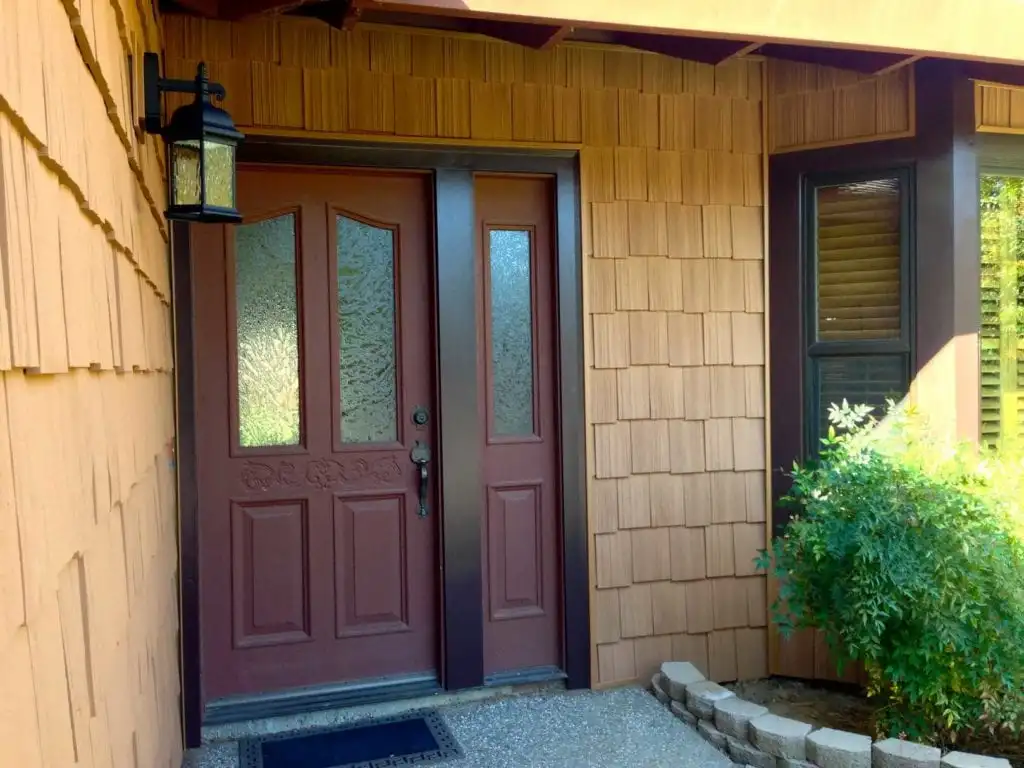 Close-up of a beige fiber cement siding wall with wood-grain texture, featuring a black metal house number "4099" and a decorative outdoor lantern casting a sharp shadow. Work by 3 Generations Improvements, Folsom.