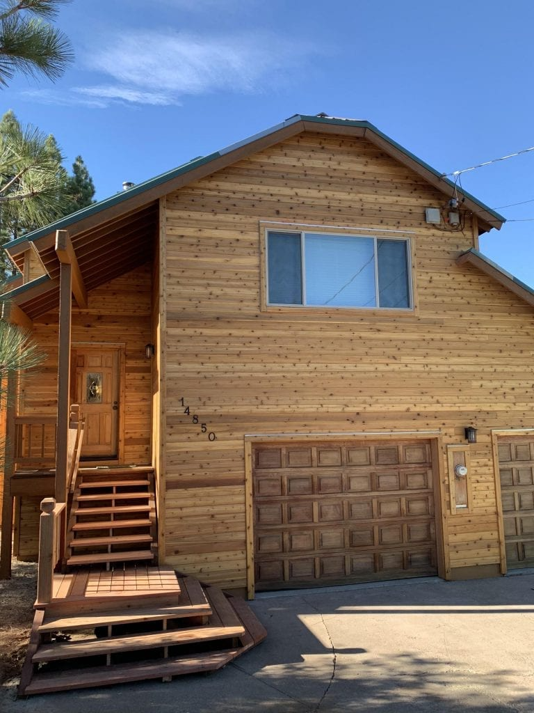 Wooden house exterior with a natural finish, featuring a wooden staircase leading to the front door and two garage doors.