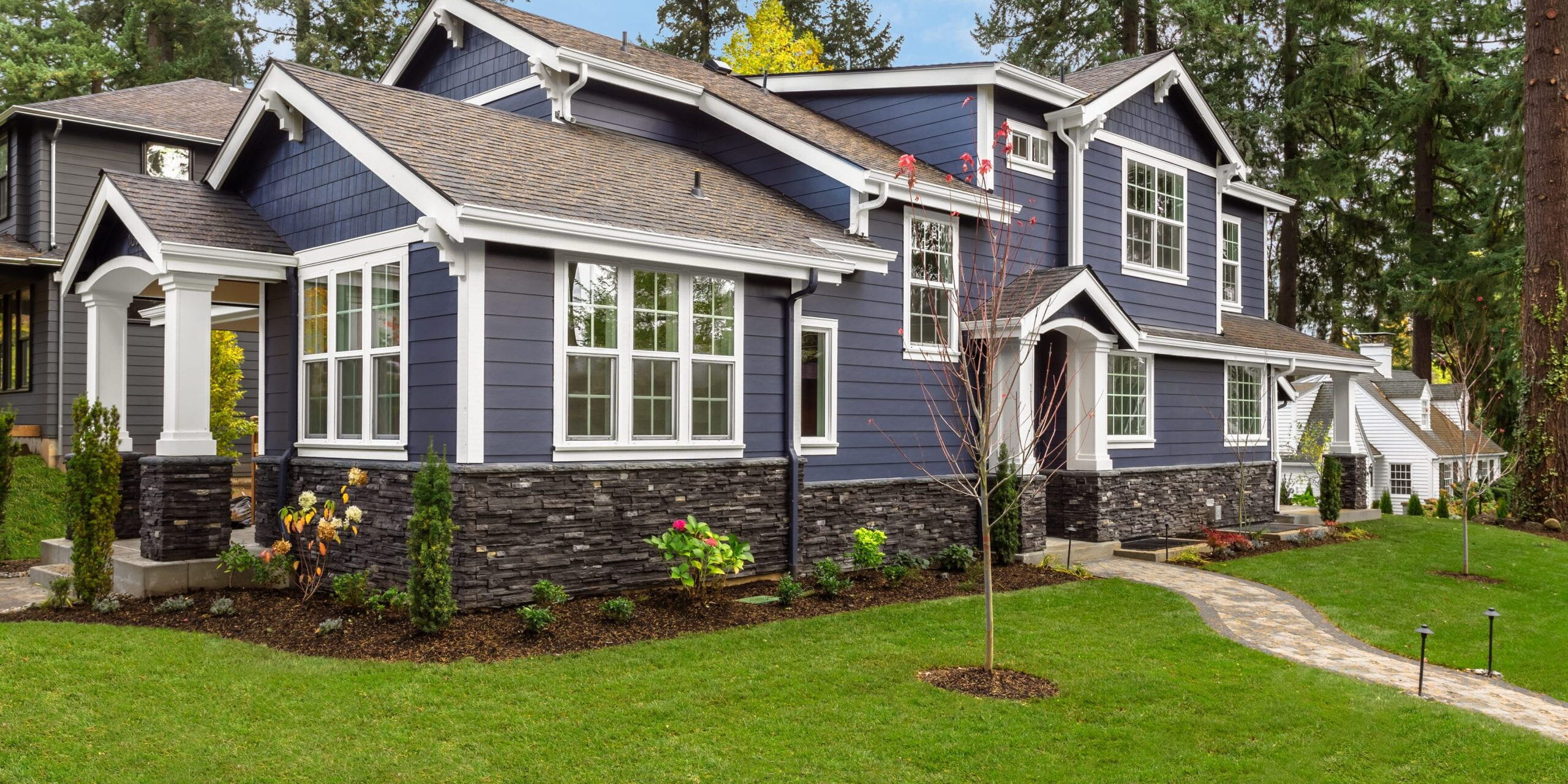 A modern two-story house with navy blue siding, white trim, and stone accents, surrounded by a well-maintained lawn and trees.