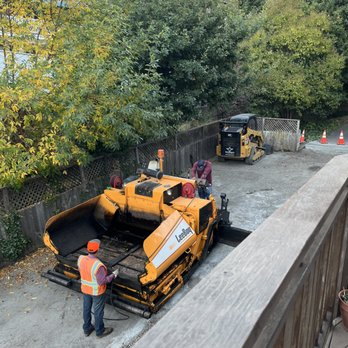 Construction workers operating paving machinery on a road in a tree-lined area.