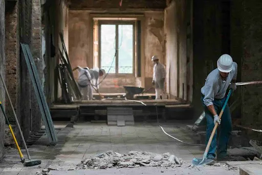 Construction workers wearing hard hats and protective gear are renovating an interior space, clearing debris and working on the structure.