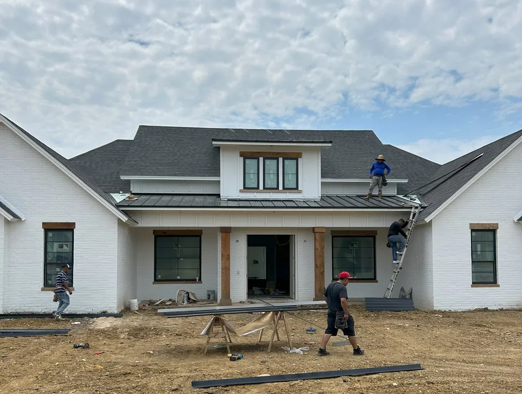 Construction crew working on the roof and exterior of a house under construction, with tools and materials visible in front of the building.