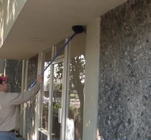 A person using a long-handled brush to clean the exterior corner of a building, representing effective pest control management by ATCO Pest Control in Petaluma.