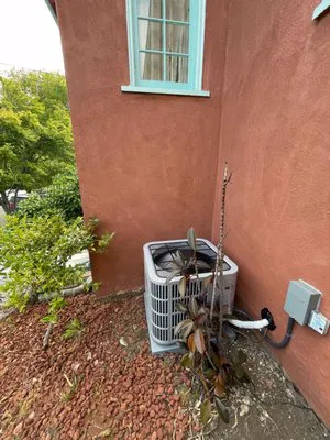 An outdoor air conditioning unit installed beside a red building, surrounded by some greenery and a small bush.