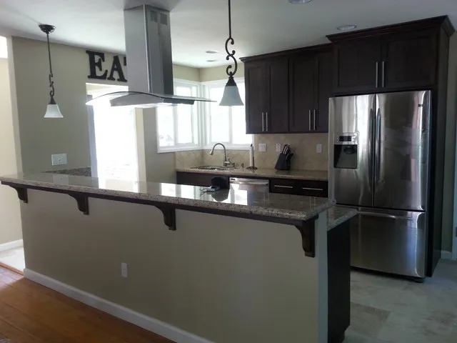 A modern kitchen interior with a granite countertop, stainless steel appliances, dark wood cabinetry, pendant lighting, and an island area.