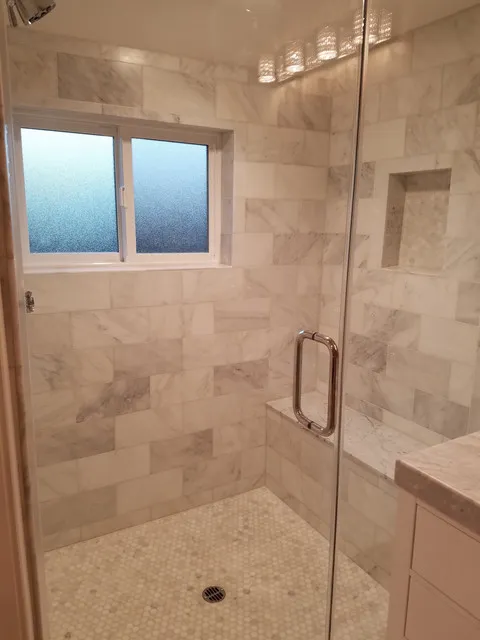 Marble-tiled shower with a glass door, built-in bench, and a frosted window designed by A Ramos Construction, Inc. in Brentwood.