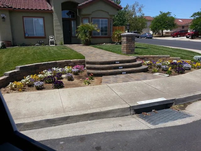 A well-maintained front yard in Brentwood featuring a brick walkway and steps leading to a house, with a neatly trimmed lawn and colorful flower beds on either side.