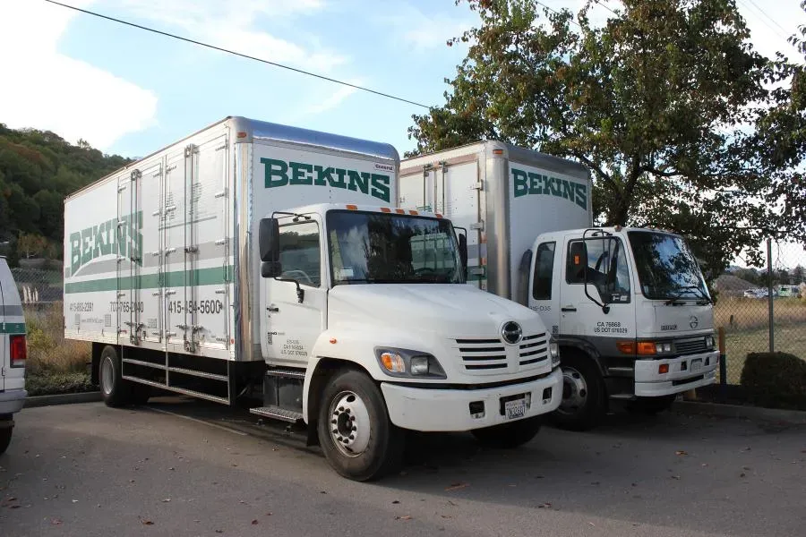 Two white moving trucks from the company Bekins parked in a lot, surrounded by trees and a chain-link fence.