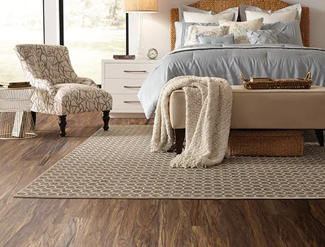A stylish bedroom with a bed featuring blue bedding, a patterned chair, and a patterned area rug on wooden floors, showcasing products from Abbey Carpets by Fashion Floors in Antioch.