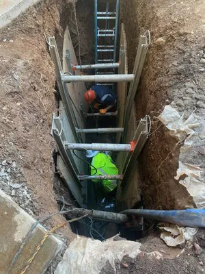 Workers in a trench equipped with safety gear and equipment, performing plumbing work.