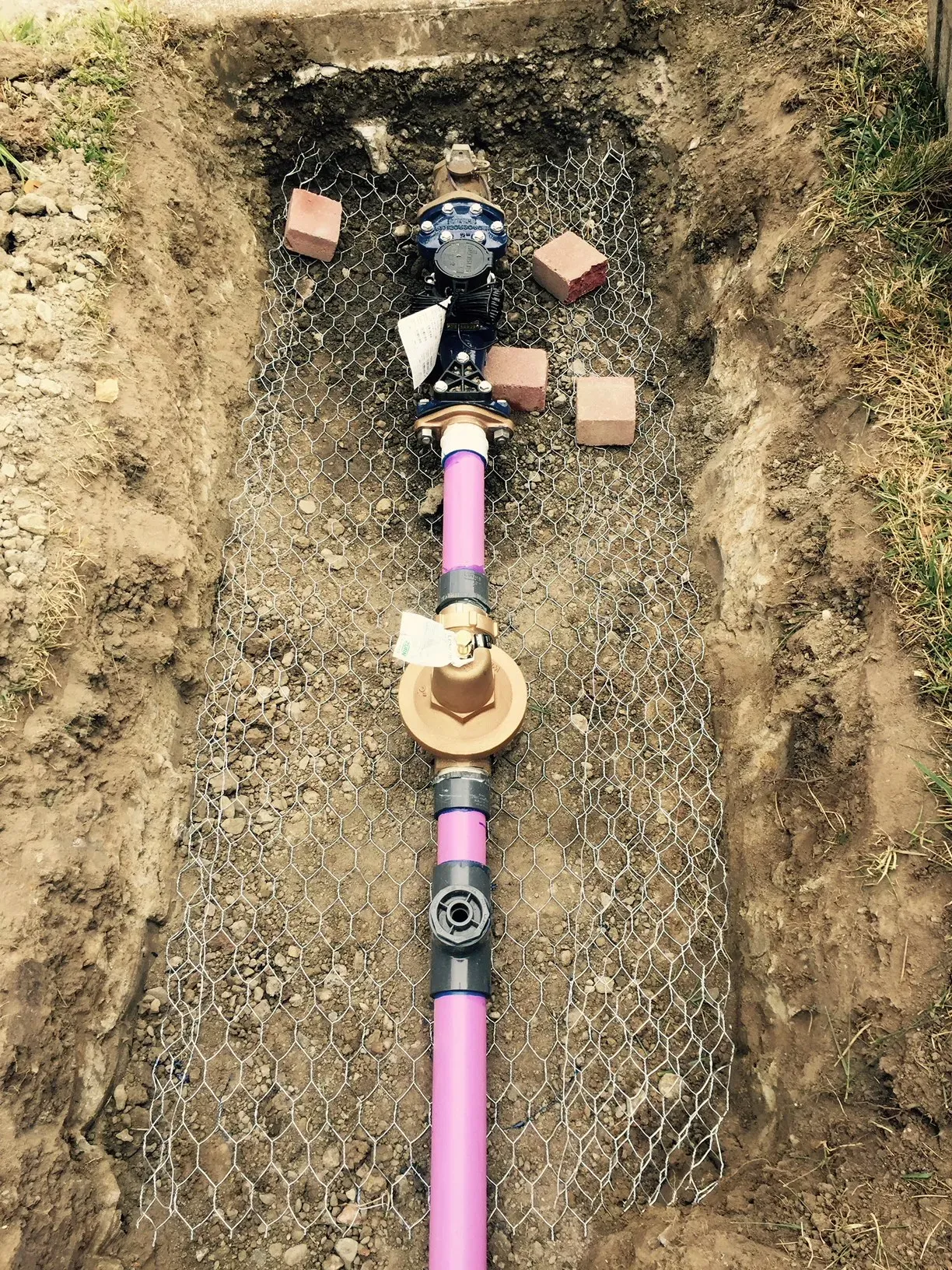 Image of a newly installed purple plumbing pipe with a valve system, surrounded by a wire mesh and bricks, in a dug trench.