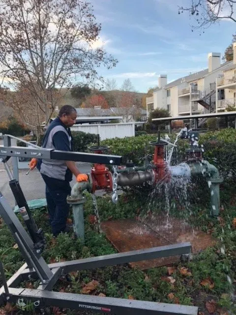 "A worker from AbsolutePlumbingandDrain in Concord fixing an outdoor plumbing valve, with water spraying from the pipe."