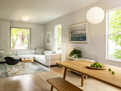 A modern living and dining room with light wood flooring, a white couch, wooden table, and large windows displaying lush greenery outside.