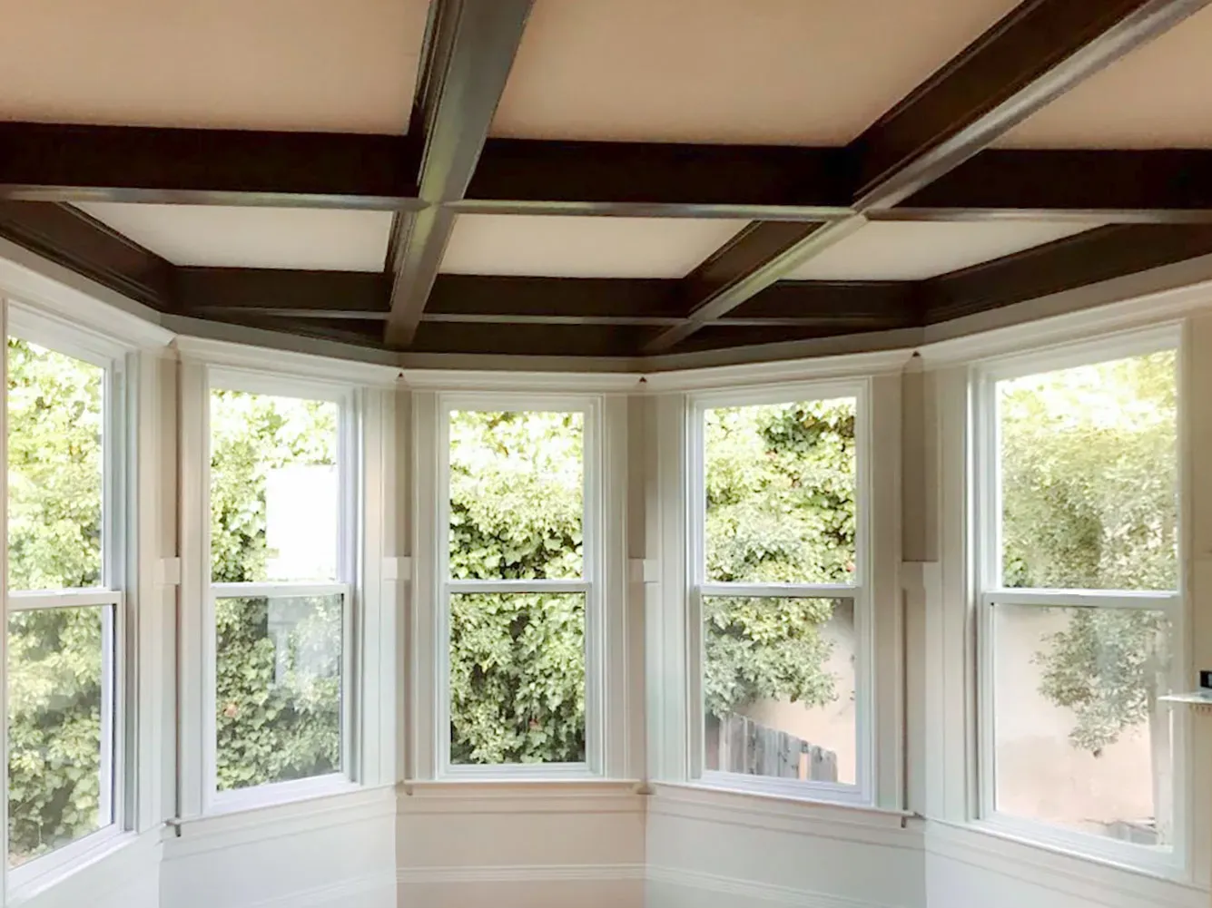 A bay window with multiple clear glass panels showcasing a lush green outdoor view, featuring a coffered ceiling above.