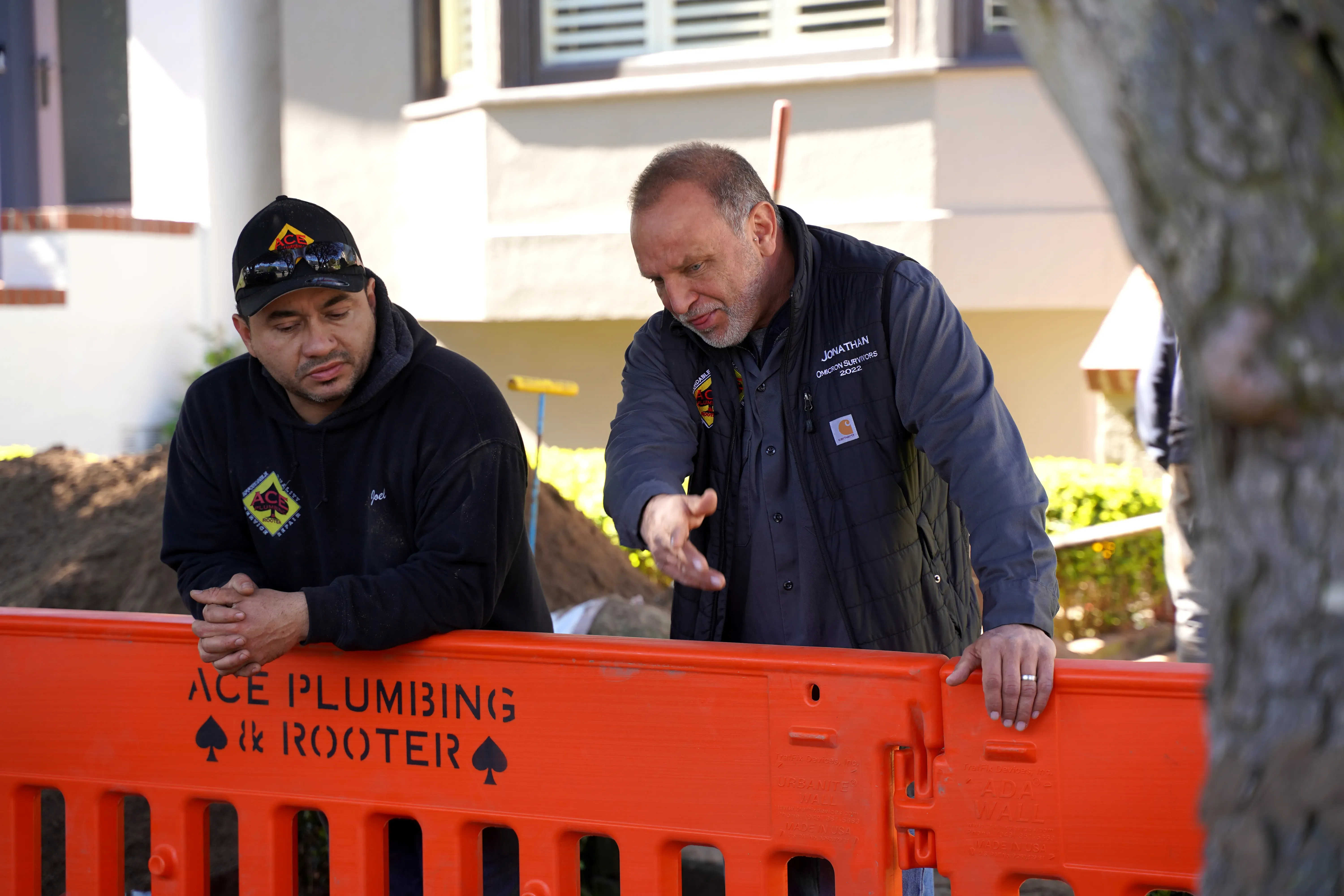 Two men in Ace Plumbing and Rooter uniforms stand behind an orange barrier in San Francisco, engaging in a focused discussion.
