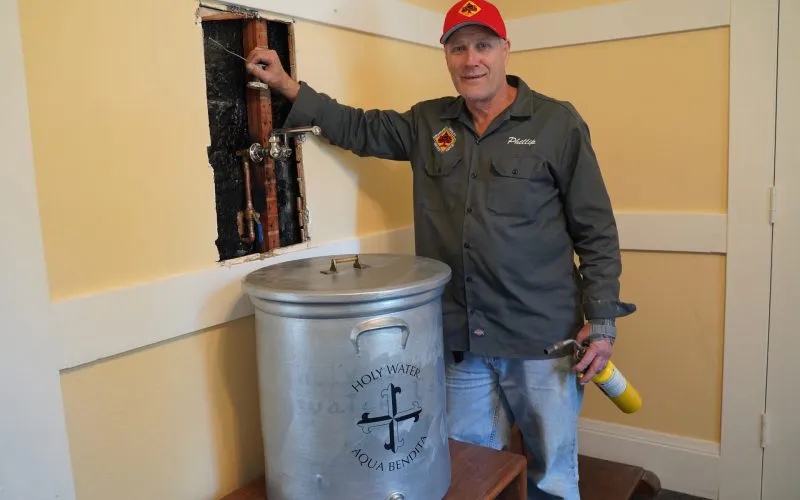 A plumber from Ace Plumbing and Rooter in San Francisco stands next to a large container labeled "Holy Water Aqua Benedicta," with plumbing pipes exposed in the wall behind him.