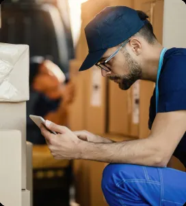 A worker in blue uniform and cap, focused on a digital tablet, surrounded by stacked boxes near a vehicle, symbolizing efficiency at Ace's Garage Door Repair & Installation in Hayward.