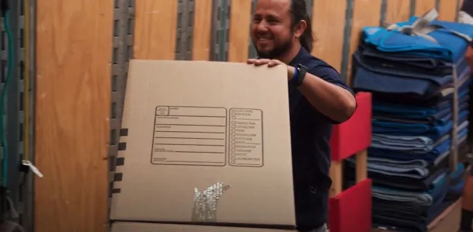 A person smiling and carrying a large cardboard box in a storage facility with shelves and packing materials nearby.