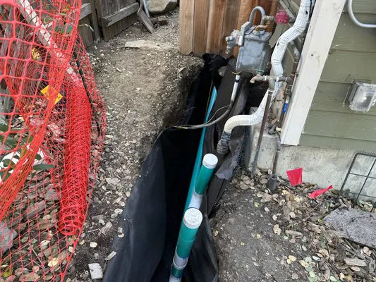 A construction site showing a trench with pipes next to a building wall, featuring utility lines and a red safety fence.