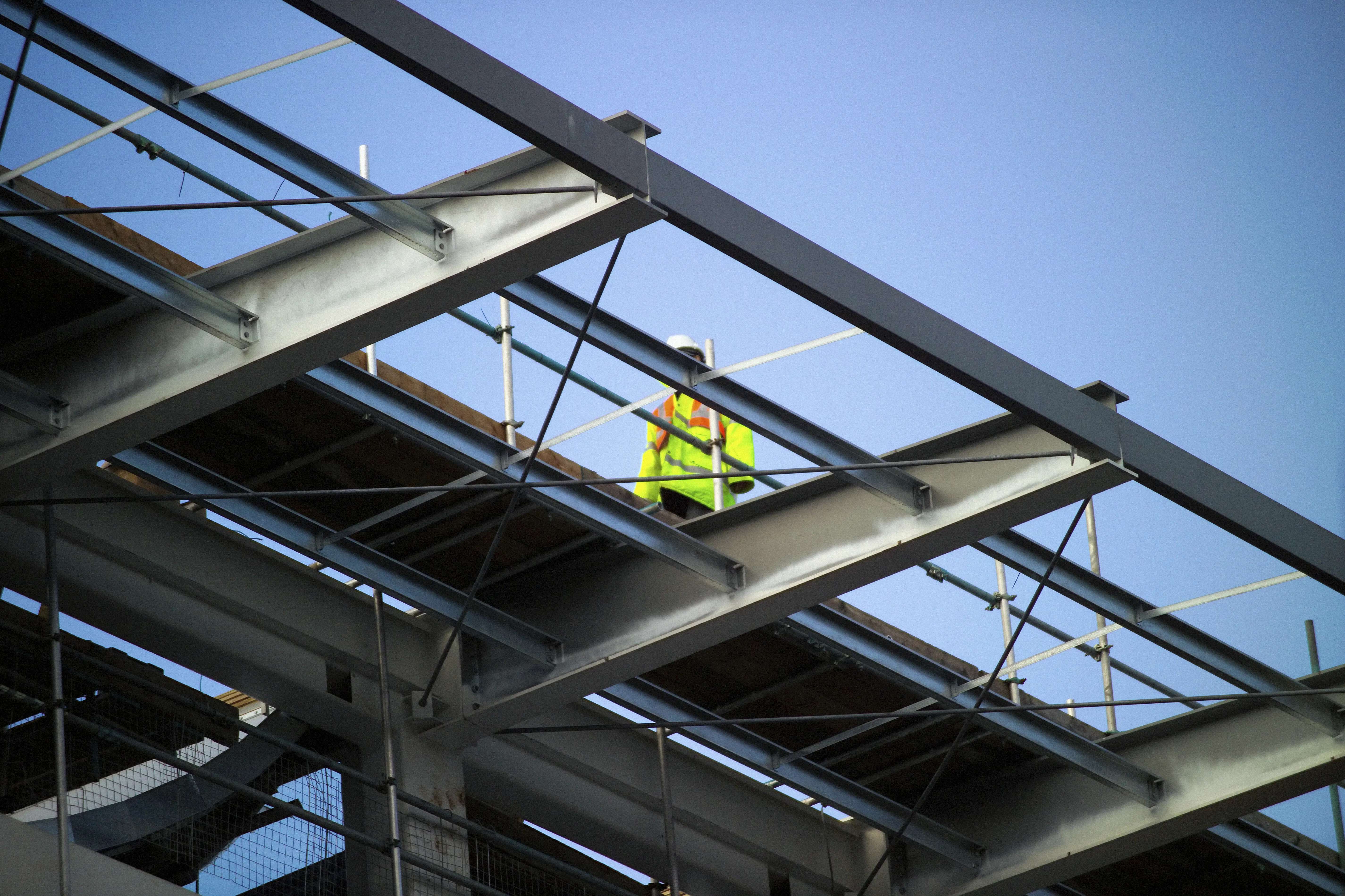A construction worker standing on a steel framework, wearing high-visibility clothing and a hard hat, against a clear blue sky background.