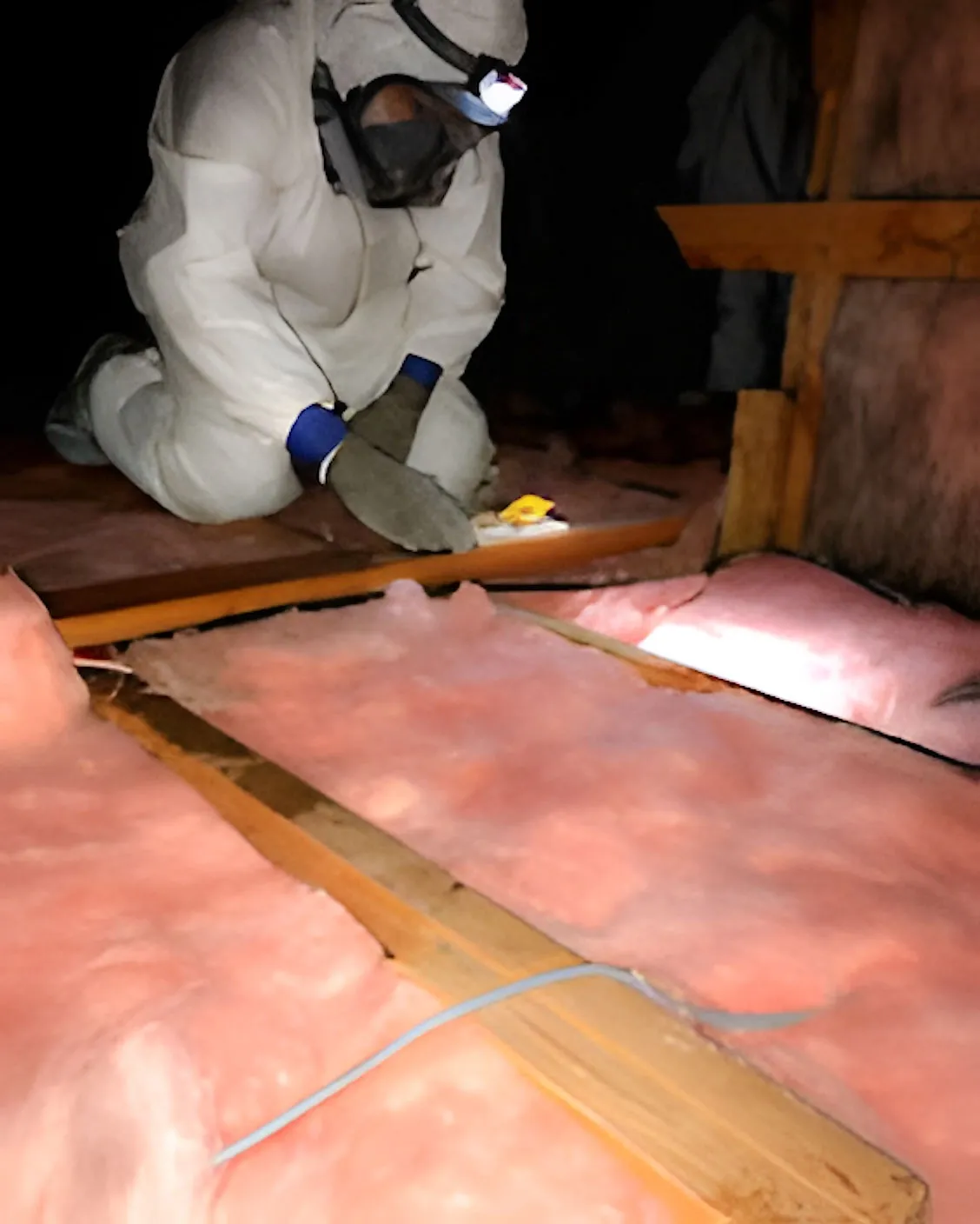 A person in protective gear is carefully working on attic insulation, surrounded by pink insulation material, emphasizing safety and precision.