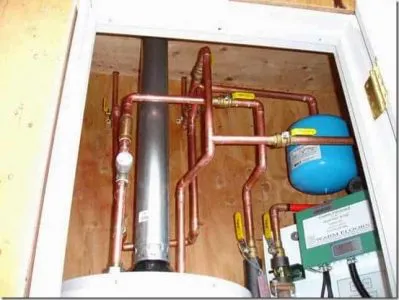 Interior view of a plumbing system with copper pipes and a blue water tank installed in a wooden enclosure.
