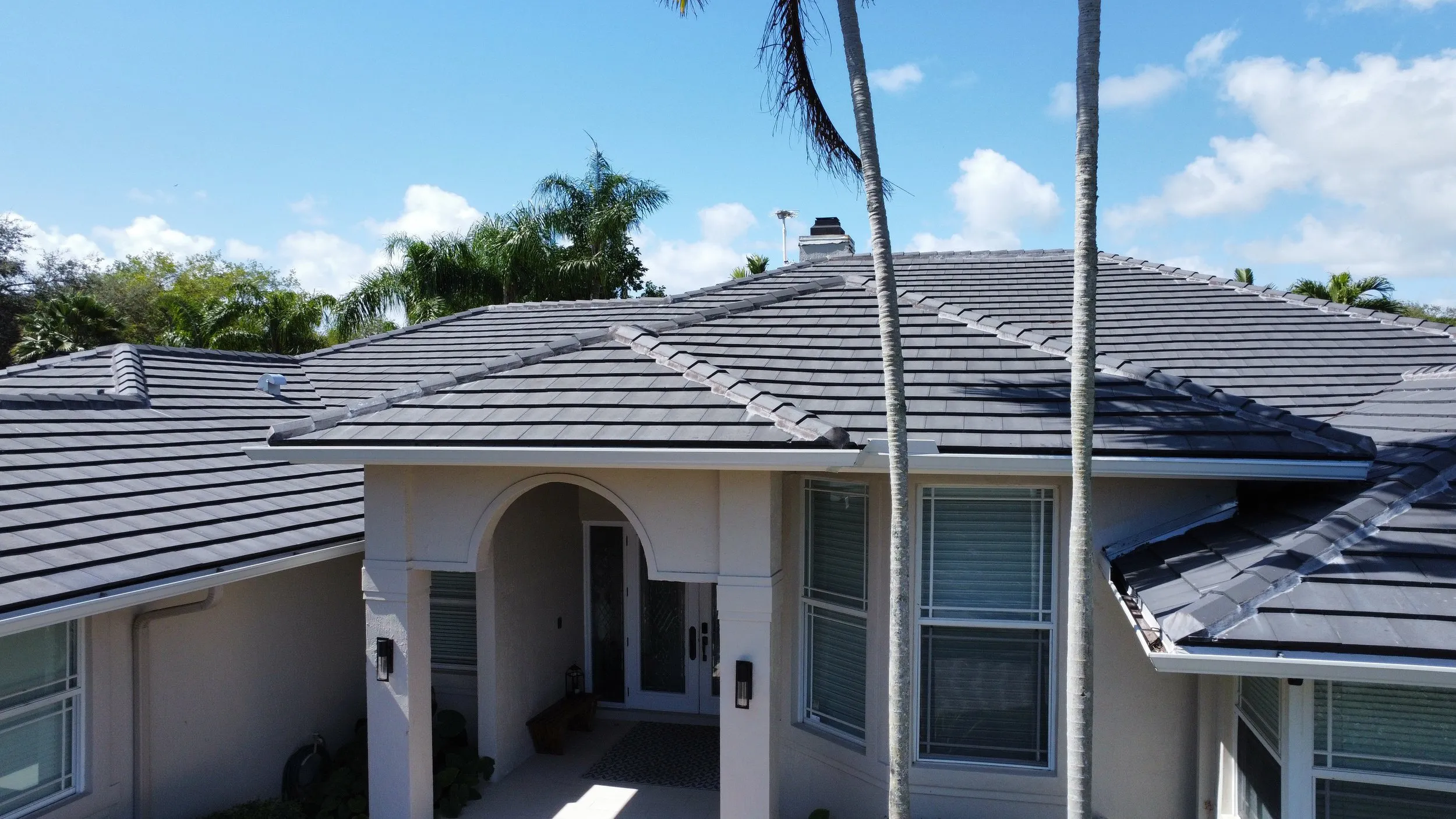 "A modern house with a newly installed gray tile roof by Alonso Roofing in Hialeah, surrounded by palm trees and a clear blue sky."