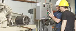Two workers in safety gear examine a control panel next to industrial equipment in a facility.