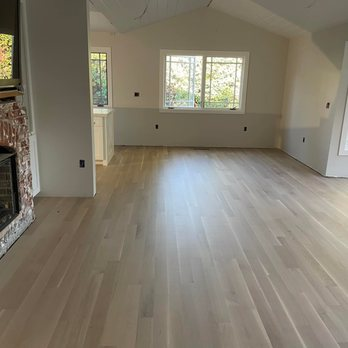 A room with freshly installed light wood flooring by Alva Flooring Inc., featuring a fireplace on the left and large windows providing natural light in Oakland.