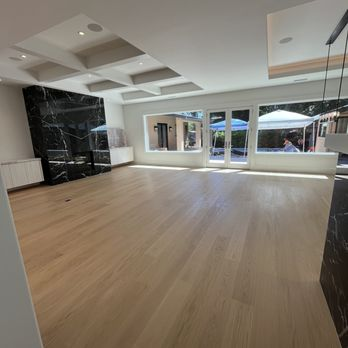 Spacious room with light wood flooring installed by Alva Flooring Inc. in Oakland, featuring large windows and a black marble accent wall.