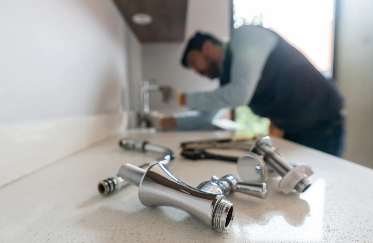 Close-up of plumbing parts on a countertop with a person working on a kitchen sink in the background.