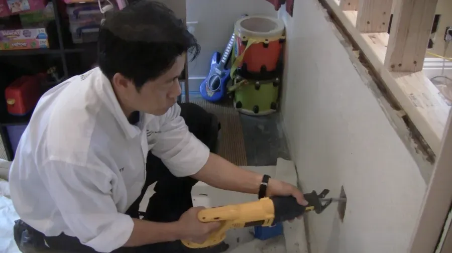 Technician using a power tool to cut into a wall, with various toys and instruments in the background.