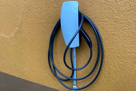 An electric vehicle charging station mounted on a textured yellow wall with a coiled black cable hanging on it.