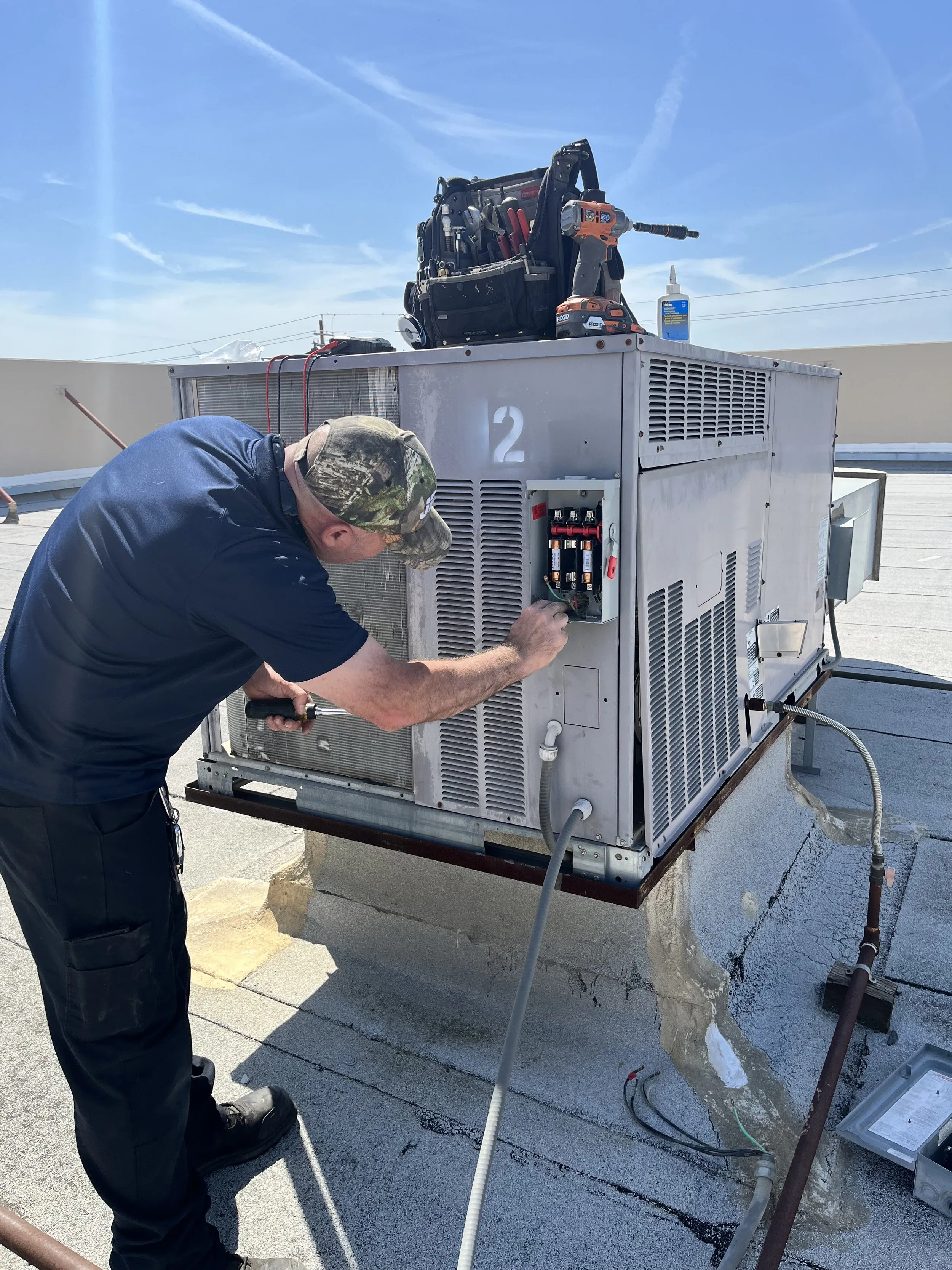 Technician from Art's Heating & Cooling performing maintenance on a rooftop HVAC unit in Pleasanton, with tools visible beside him.