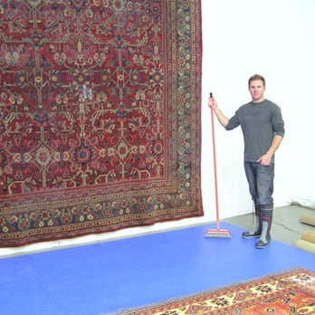 A person stands next to an ornate red rug hanging on a wall, holding a cleaning tool in a professional setting.