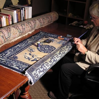A person carefully working on restoring a blue and beige rug on a wooden table, with tools and thread visible.