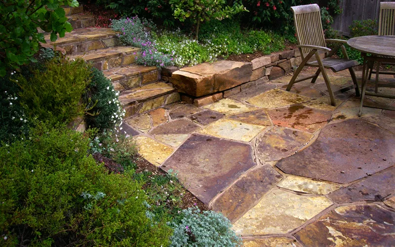 A well-designed stone patio featuring diverse stone slabs, bordered by lush greenery and flowering plants, with wooden chairs and a table, designed by Artisans Landscape Inc in San Francisco.