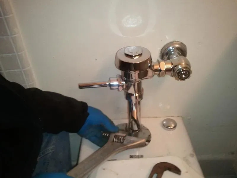 A plumber in blue gloves using a wrench to adjust a toilet flushometer, showcasing professional plumbing work by Atlas Plumbing & Rooter in San Francisco.