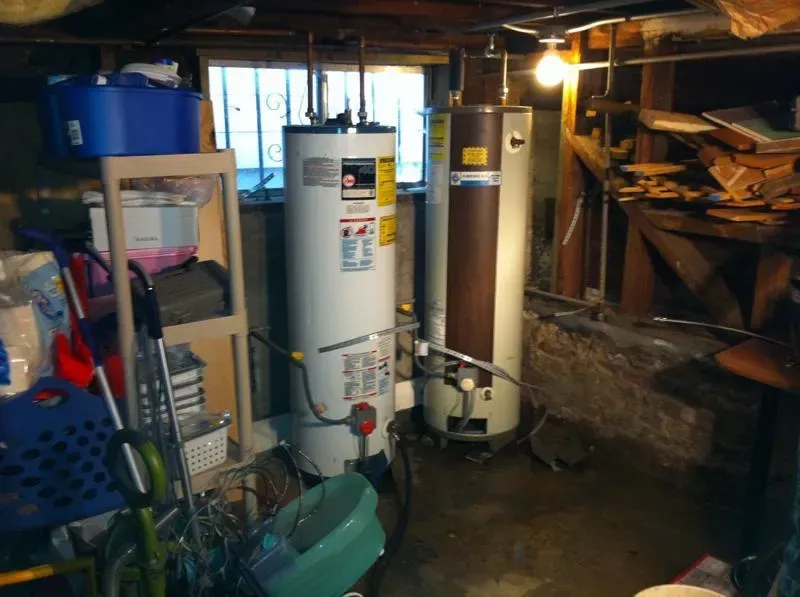 A basement with two water heaters, surrounded by storage items and wooden shelves.
