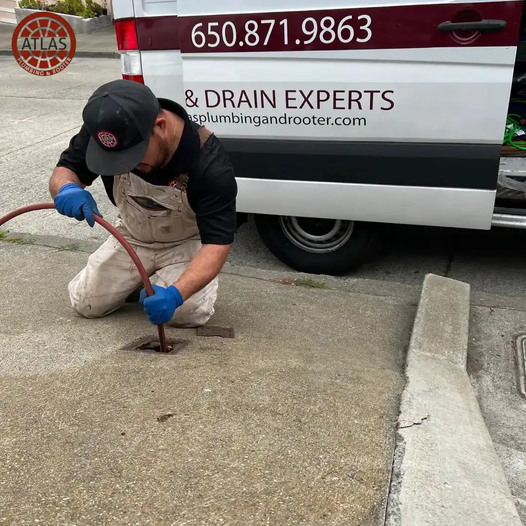 "A technician from Atlas Plumbing & Rooter works on a drain in San Francisco, kneeling on the pavement next to a service van."