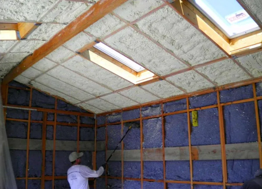 Worker in an attic applying insulation to walls and ceiling, with skylights present.