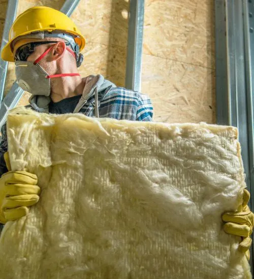 Worker wearing protective gear holding a sheet of insulation material in an attic space.