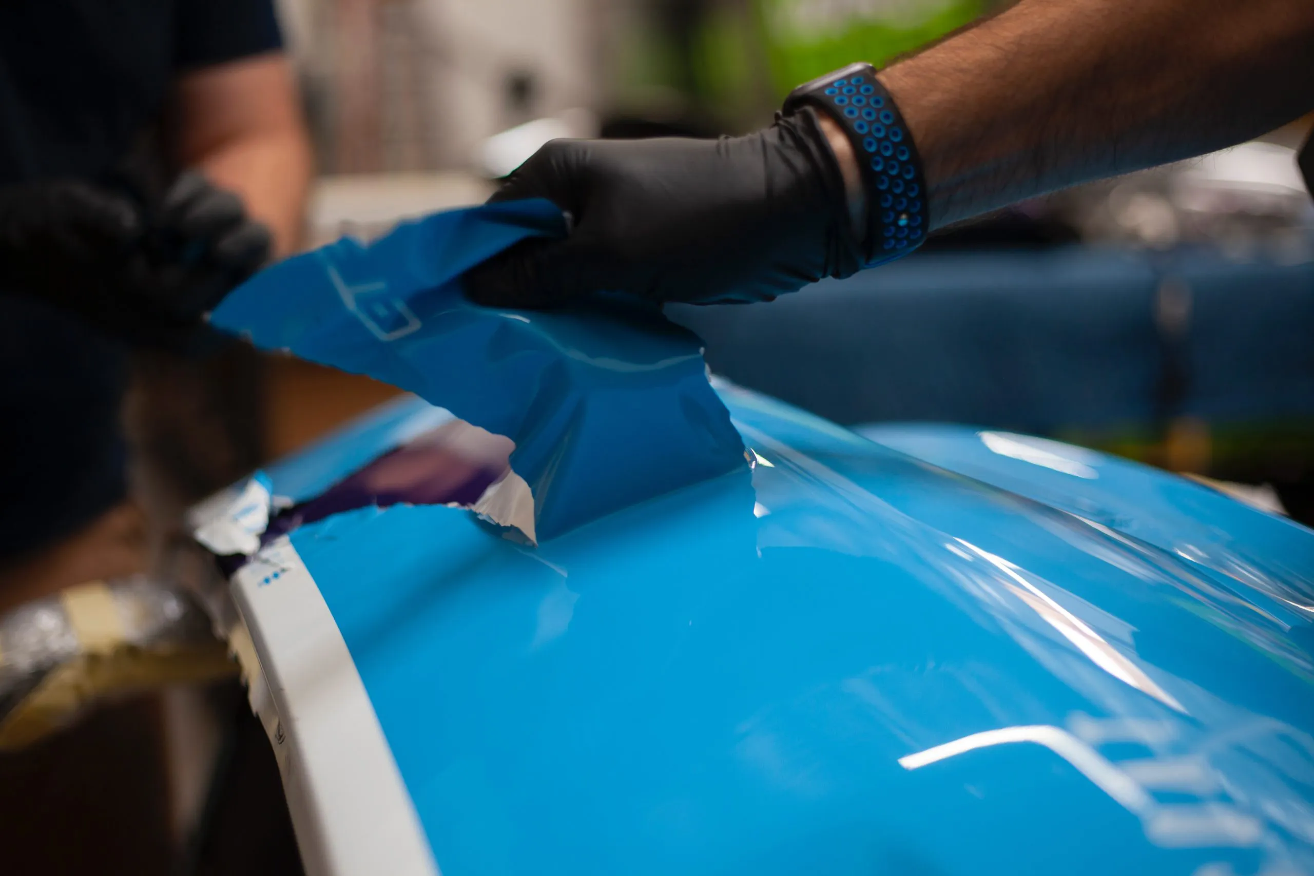 A technician wearing a glove carefully applies blue vinyl wrap to a car's surface at AutoShield Collision in Santa Clara.