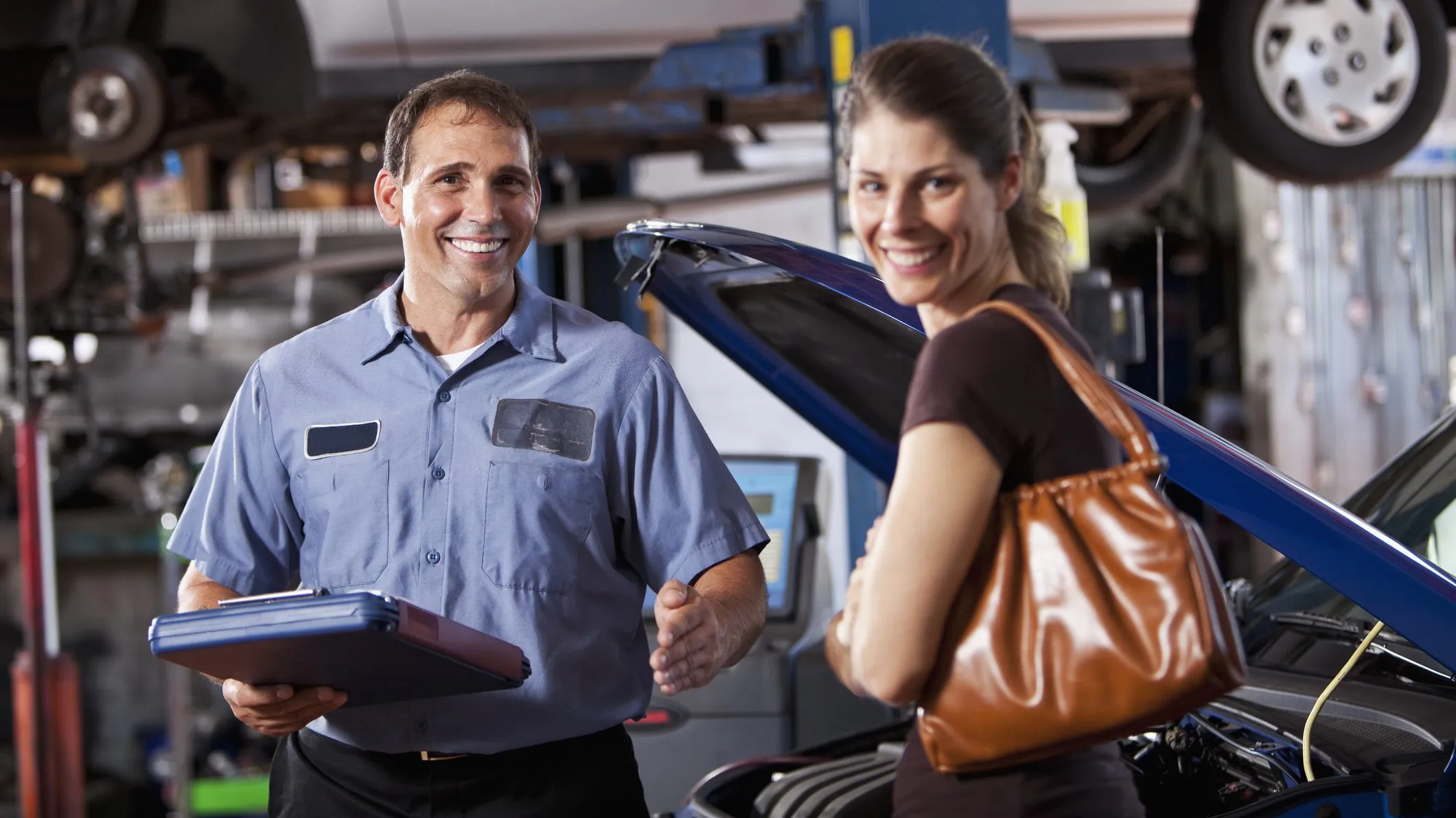 A mechanic holding a clipboard smiles at a customer near a car with its hood open, at B2 Perfection Auto Body in Sunnyvale.