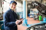 Mechanic in a blue uniform with a clipboard standing next to a car with its hood open inside an auto repair shop.