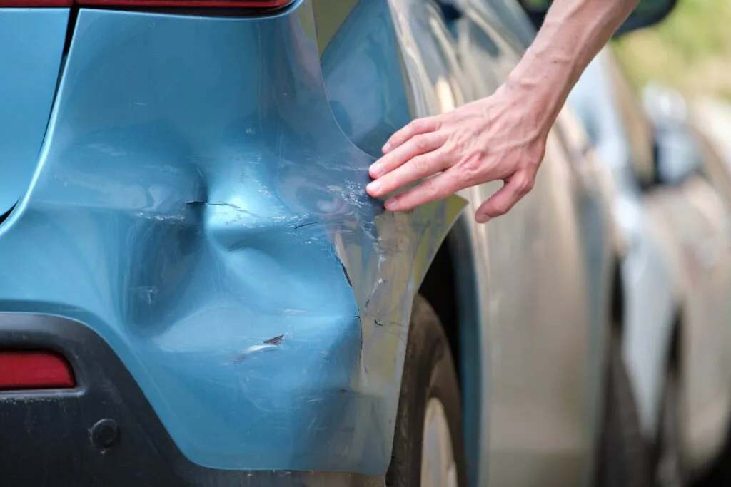 A hand inspecting a dent and scrape on the rear side of a blue car.