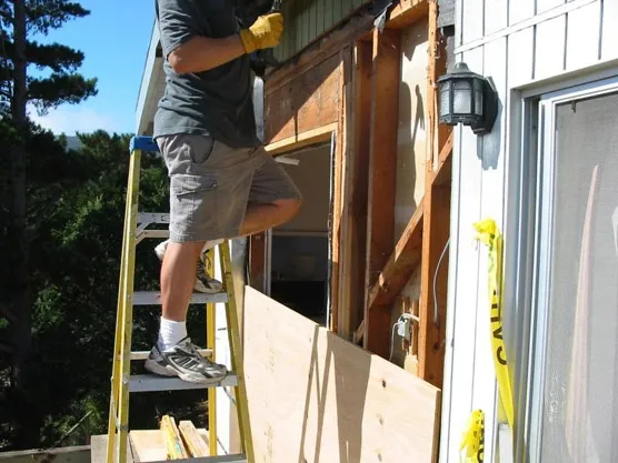 A construction worker from BB & B Builders Inc. stands on a ladder next to a partially renovated exterior wall of a house in Mendocino, wearing gloves and holding onto the structure.