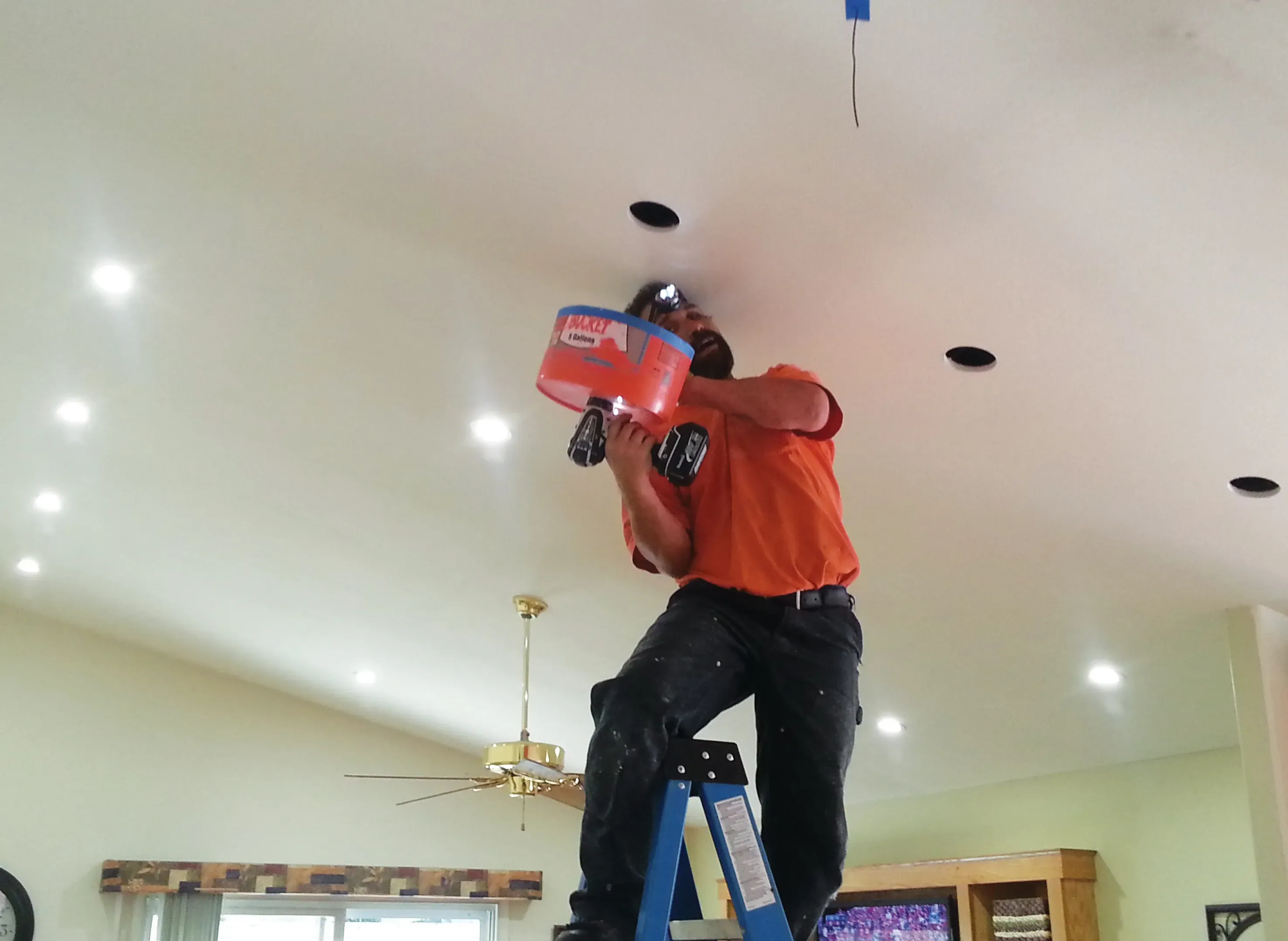 An electrician in an orange shirt from B. Henry's Quality Electric Inc. stands on a ladder in a room in Petaluma, installing lighting fixtures in the ceiling.