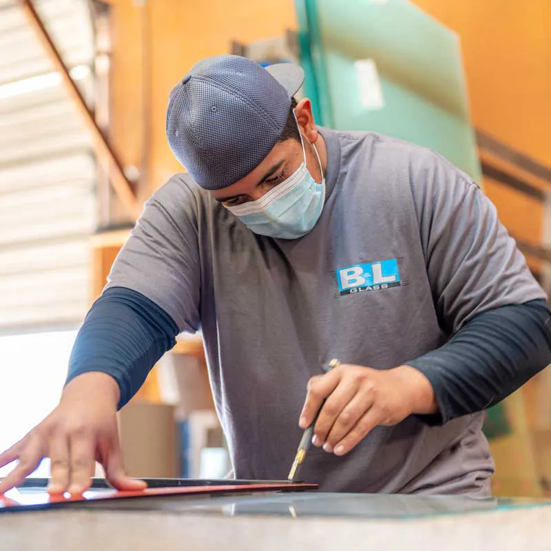 A worker from B & L Glass in Santa Rosa wearing a grey uniform and face mask uses a tool to cut glass in a workshop.